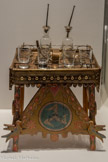 <center>Table de marchand de sirop ambulant</center>Début du 20e siècle Palerme, Italie
Verre incolore, bois peint, métal
Collection du Muséum national d'histoire naturelle
Cet objet est un modèle réduit d’une table d’acquaiolo, marchand ambulant d’eau aromatisée. En Sicile, cette pratique est attestée dès le 18e siècle. Le sirop était conservé dans les bouteilles et les verres étaient utilisés pour servir les passants.
La table est décorée du triskele, symbole de la Sicile.