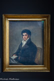 <center>Le musée Goya.</center>Francisco de GOYA Y LUCIENTES
(FUENDETODOS, 1746 - BORDEAUX, 1828)
Portrait de Francisco del Mazo
VERS 1815-1820 HUILE SUR TOILE
Né en 1772, Francisco del Mazo était « agente de Casa » au sein de la maison du duc d’Albe en 1793.
En 1804-1805, il fait partie de la Junta de Gobierno de la fameuse banque de Saint-Charles, future banque d’Espagne fondée par François Cabarrus en 1782. En 1815, il occupe enfin le poste de premier comptable du Mont de Pitié des Caballejos Hijosdalgos de Madrid tout en étant lié à l’Inquisition. Ce portrait a été réalisé à l’occasion d’une promotion sociale, peut-être un anoblissement. En 1805, Goya écrivait à un ami qu’il fallait payer plus cher lorsque les mains du modèle, morceau de bravoure, devaient être apparentes. Francisco del Mazo n’a pas dû payer le prix le plus élevé, qui se situait entre 5 000 et 15 000 réaux pour des portraits en buste ou en pied : en effet ses mains n’apparaissent pas vraiment, l’une étant cachée par le billet et l’autre glissée dans le gilet. Pourtant la qualité de l’œuvre ne s’en ressent pas et la personnalité énergique et avisée du modèle est perceptible. Goya ne s’embarrasse d’aucun artifice, en particulier pour l’éclairage : la lumière est juste figurée d’un trait rapide d’ocre jaune sur le dossier du fauteuil. Le fond uni, quasi systématique après 1815, renforce la présence humaine tout comme chez les modèles de Velâzquez. Identifiée grâce â l’inscription du billet tenu dans la main droite, l’œuvre est datée entre 1815-1820 en raison de la redingote à col haut dite « à l’anglaise » qui était à la mode après la chuta de l’Empire Napoléonien.